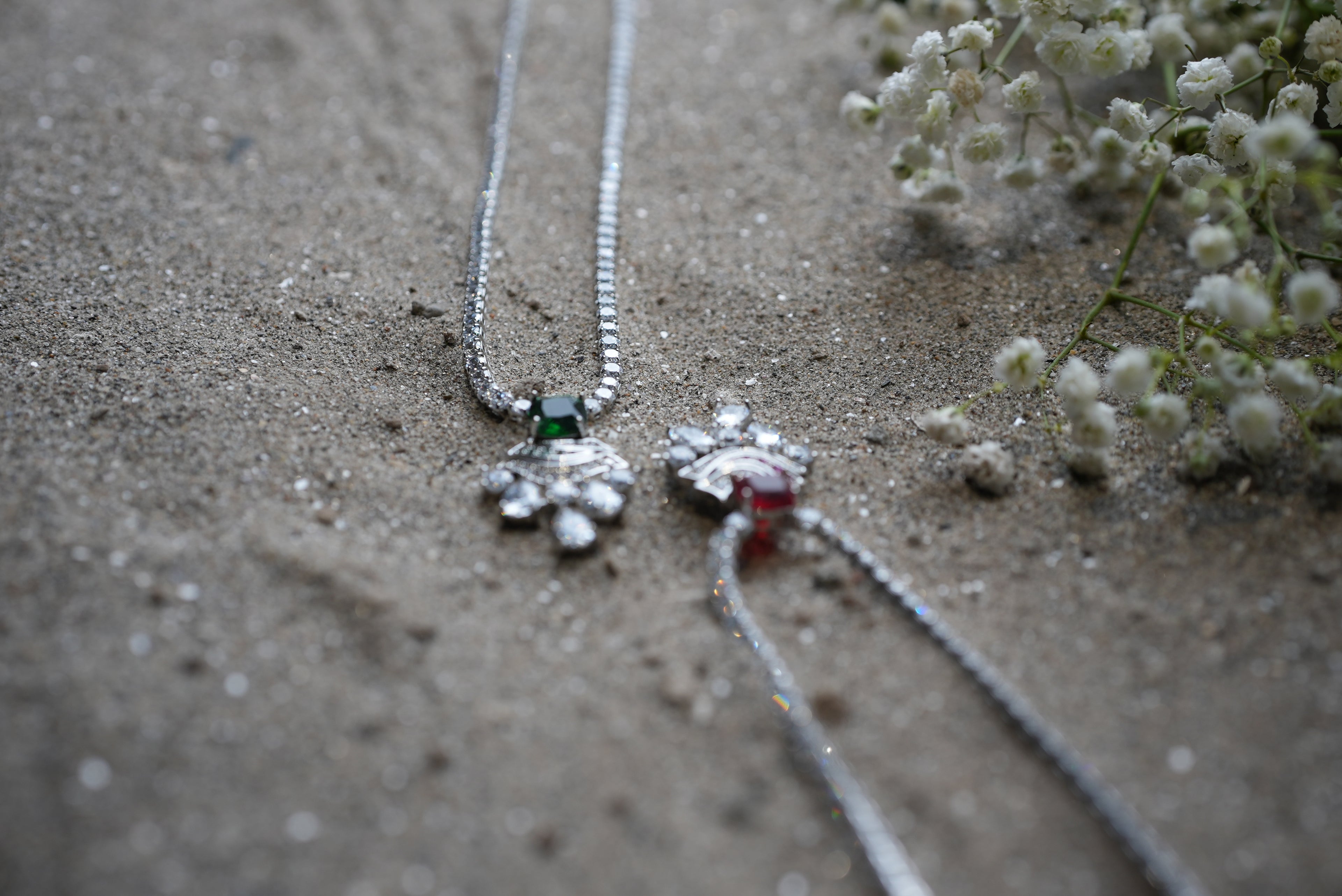 Two silver necklaces with floral pendants on a sandy surface.