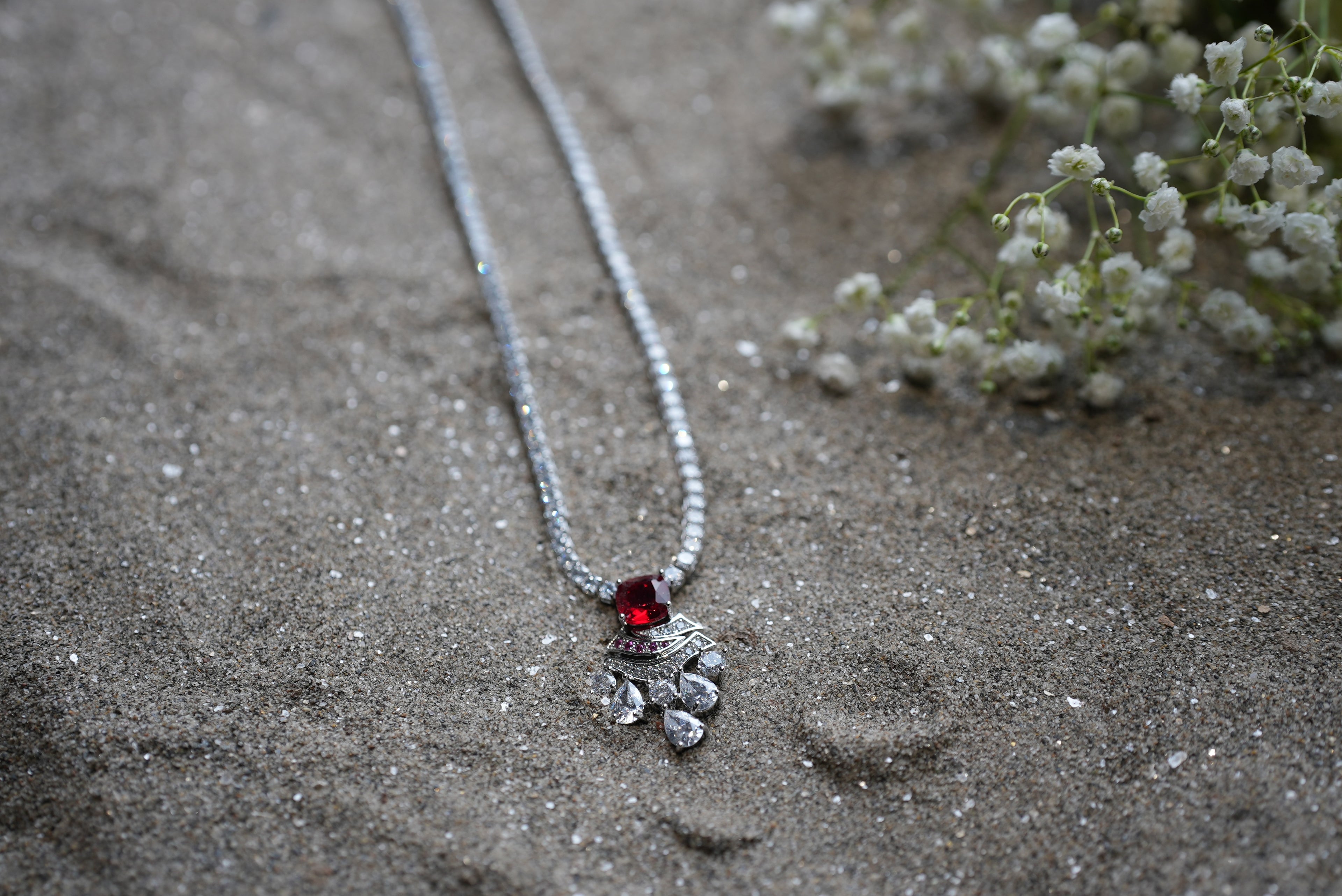 Silver necklace with a red gemstone pendant on sandy ground with small white flowers.