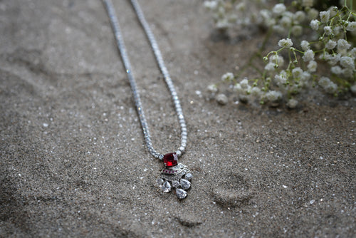 Silver necklace with a red gemstone pendant on sandy ground with small white flowers.